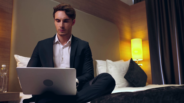 молодой серьезный бизнесмен, сидящий на кровати и с ноутбуком в номере отеля
  - Кадры, видео