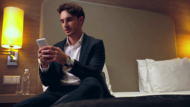 χαμογελαστός επιχειρηματίας που κάθεται στο κρεβάτι και συνομιλεί στο smartphone στο δωμάτιο του ξενοδοχείου - Πλάνα, βίντεο