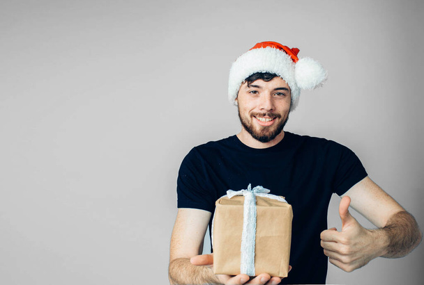 Νεαρός γενειοφόρος απομονωμένος στο βάθος. Ένας τύπος με μαύρο πουκάμισο και εορταστικό κόκκινο χριστουγεννιάτικο καπέλο που ποζάρει στην κάμερα. Κρατήστε τον μεγάλο αντίχειρα επάνω και το κιβώτιο του δώρου ή παρόν στο χέρι. - Φωτογραφία, εικόνα