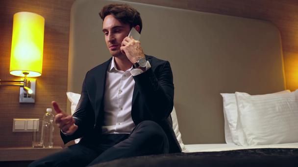 uśmiechnięty biznesmen siedzący na łóżku i rozmawiający na smartfonie w pokoju hotelowym - Materiał filmowy, wideo