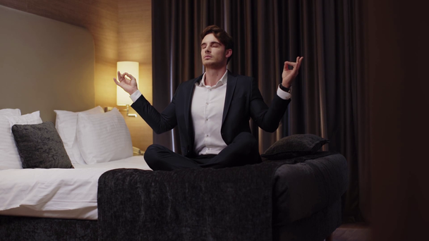 young businessman meditating in lotus pose on bed in hotel room - Felvétel, videó