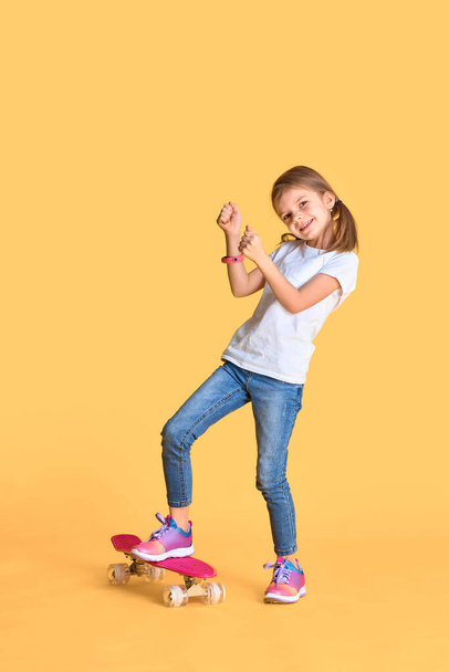 Stylish funny girl wearing white t-shirt, blue jeans and sneakers, standing on skateboard over yellow background - Φωτογραφία, εικόνα