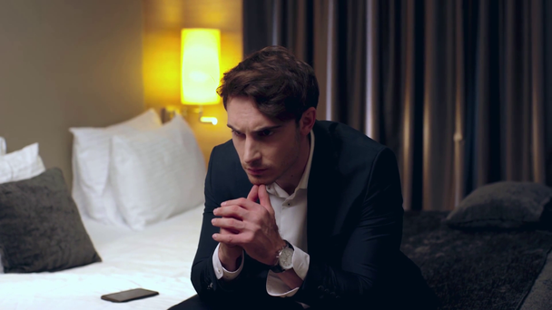 zdenerwowany biznesmen siedzi na łóżku z zaciśniętymi rękami w pokoju hotelowym - Materiał filmowy, wideo