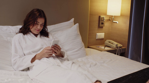 happy girl in bathrobe chatting on smartphone while resting in bed - Footage, Video