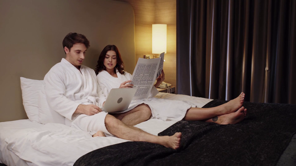 man met laptop en vrouw die de krant leest terwijl hij rust in de hotelkamer - Video