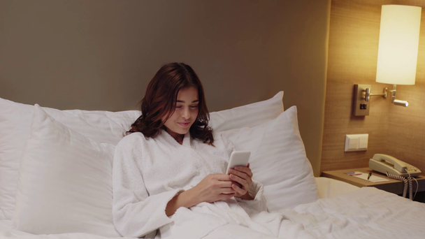 Bornozlu gülümseyen kız yatakta dinlenirken akıllı telefondan sohbet ediyor. - Video, Çekim