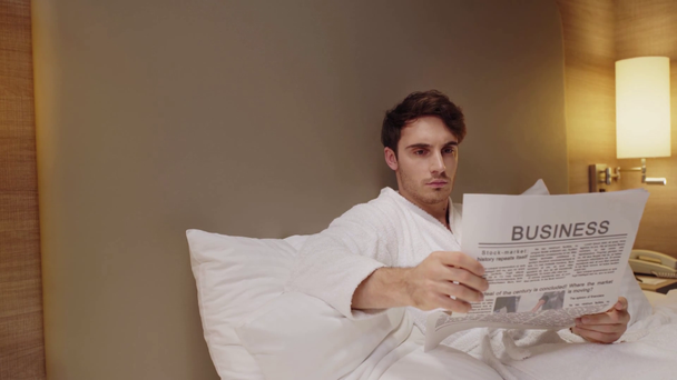 joven, hombre concentrado leyendo el periódico de negocios mientras descansa en la cama
  - Imágenes, Vídeo