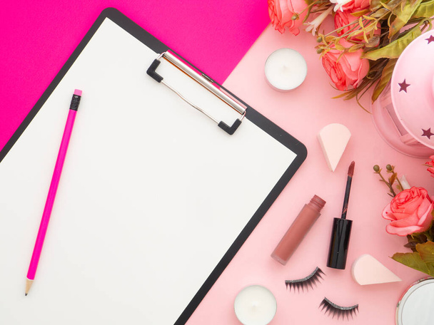 Clipboard and makeup tools and cosmetic products and roses flowers around it isolated on a pink and peach color background. Flat lay, top view copy space. Feminine beauty blogger workplace concept. - Foto, Imagem