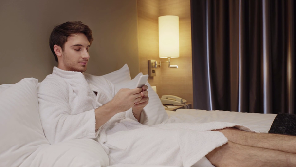 jonge geconcentreerde man glimlachen tijdens het rusten in bed en chatten op de smartphone - Video