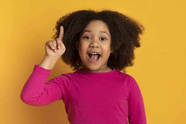 Ιδέα. Μικρό μαύρο κορίτσι δείχνει το δάχτυλο πάνω από το κίτρινο φόντο - Φωτογραφία, εικόνα