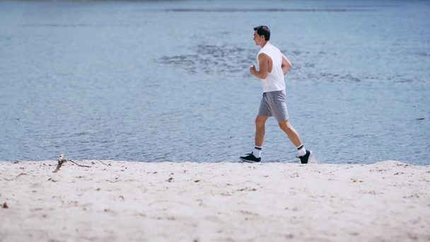 vista lateral do jovem desportista correndo ao longo da costa do rio areia
 - Filmagem, Vídeo