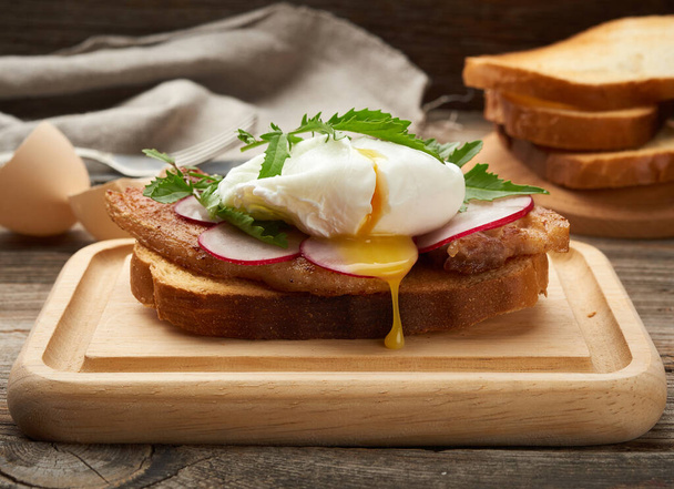sandwich sur une tranche de pain blanc grillé aux œufs pochés, feuilles vertes de roquette et radis, petit déjeuner matinal sur une planche de bois brun
 - Photo, image