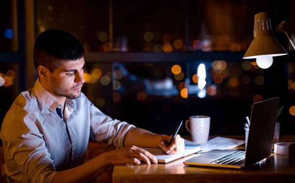 Freelancer Guy tomando notas sentado no laptop trabalhando remotamente durante a noite no Home Office
 - Foto, Imagem