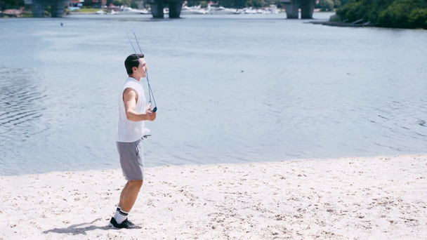 side view of young sportsman jumping with skipping rope on sand shore - Materiał filmowy, wideo