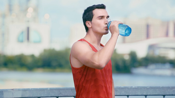 橋の上に立ってスポーツボトルから水を飲む若いスポーツマンは - 映像、動画