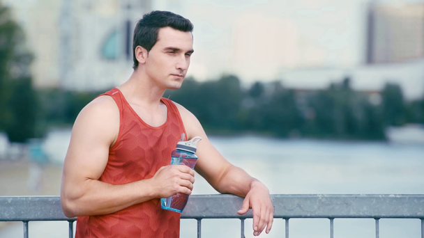 νεαρός αθλητής στέκεται στη γέφυρα και πόσιμο από αθλητικό μπουκάλι - Πλάνα, βίντεο
