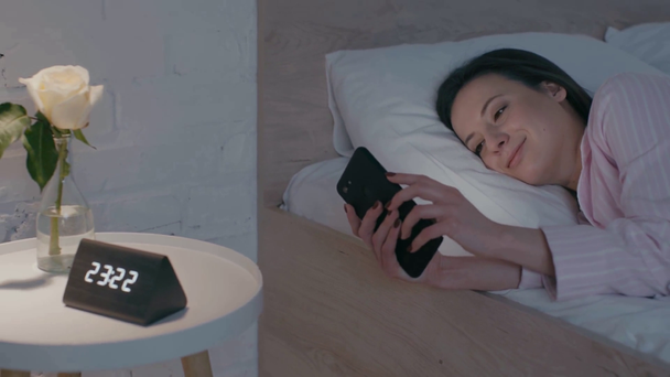 Mosolygós lány használ okostelefon közel óra és virág az éjjeliszekrényen  - Felvétel, videó