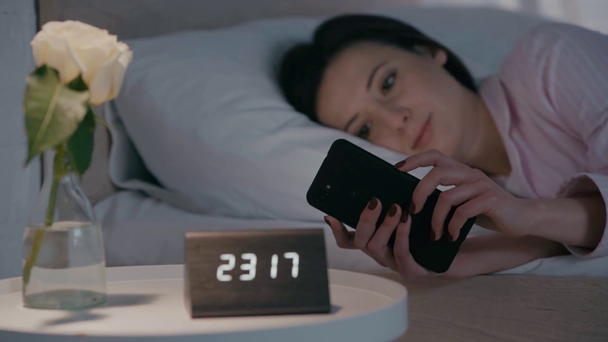 Yatakta akıllı telefon kullanan bir kızın yanındaki masada gül ve saatin seçmeli odak noktası - Video, Çekim
