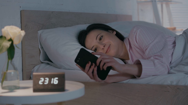 Selective focus of smiling woman using smartphone on bed at night - Imágenes, Vídeo