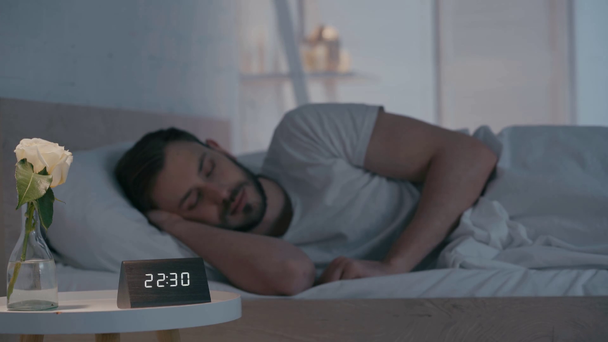Selektywne skupienie zegara i kwiatu na stole w pobliżu śpiącego mężczyzny na łóżku - Materiał filmowy, wideo
