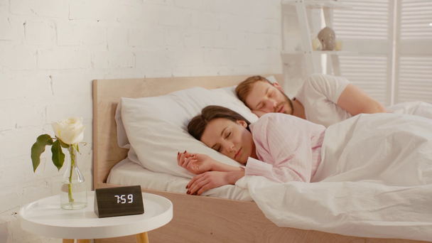 Couple sleeping on bed near clock and rose in vase on bedside table  - Filmati, video