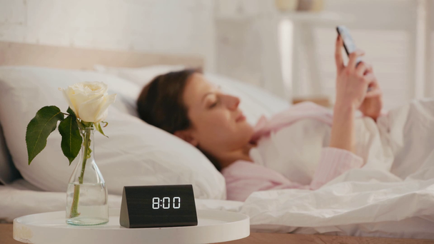 Clock and rose on table near smiling girl using smartphone on bed at morning - Footage, Video