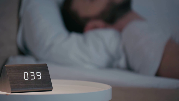 Selective focus of man looking at clock while lying on bed at night - Materiał filmowy, wideo