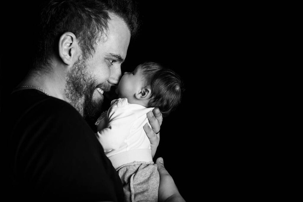 Studio portret van knappe gespierde man in zwart t-short vasthouden aan handen schattig kleine baby over zwarte achtergrond met copyspace. Familie, liefde en geluk. - Foto, afbeelding
