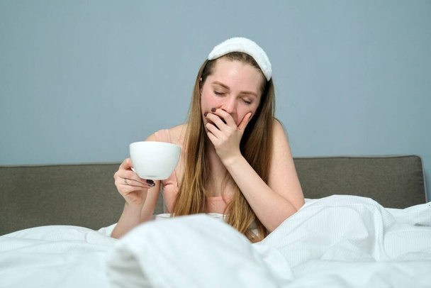 Sleepy yawning young woman in bed with cup of tea, morning, white bed, background gray wall - Fotografie, Obrázek