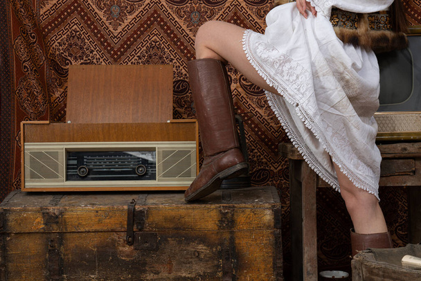 Young female in national white dress and old leather shoes puts her leg on the aged chest near old radio - Fotoğraf, Görsel