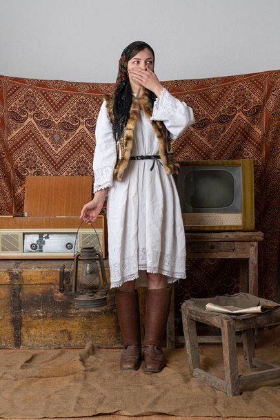 Ulusal Ukrayna elbiseli genç bir kadın eski televizyon ve radyonun yanındaki klasik odada duruyor elinde gazyağı lambası, eliyle ağzını kapatıyor ve sağ tarafına bakıyor. - Fotoğraf, Görsel