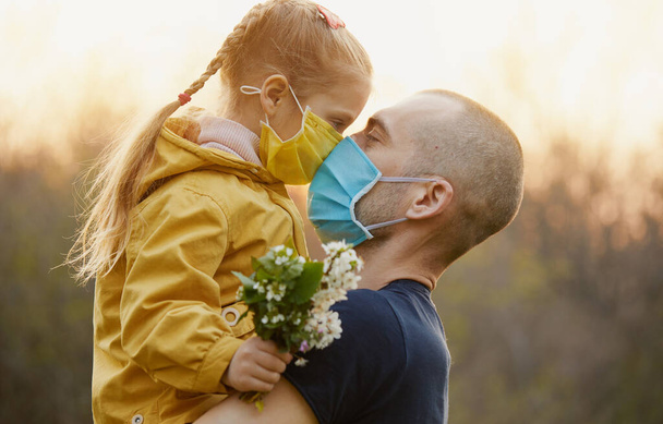 Family protection. A young father hugs his little girl daughter in the spring garden outdoors. Protective masks on faces from coronavirus infection - Foto, afbeelding