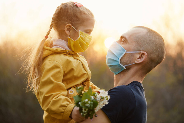 Family protection. A young father hugs his little girl daughter in the spring garden outdoors. Protective masks on faces from coronavirus infection - Photo, Image