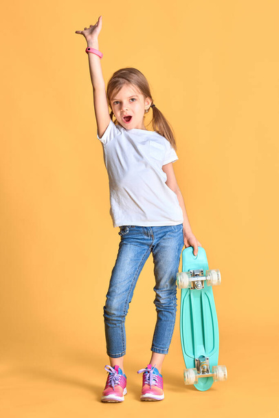 Stylish funny girl wearing white t-shirt, blue jeans and sneakers, holding skateboard over yellow background - Fotografie, Obrázek