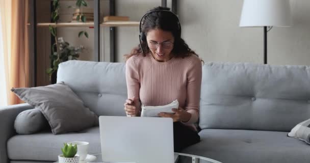 Młoda kobieta w słuchawkach dzwoniąca na laptopa w domu - Materiał filmowy, wideo