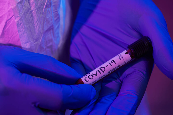 Coronavirus, Doctor holding positive covid-19 virus Blood Sample test tube. Wearing biohazard epidemic Protective mask, suit and glows neon light background - Foto, Imagen