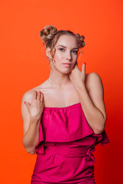 The girl in a red dress on a orange background in the studio. Blonde girl with two hair knots holding hands on shoulders, looking to the camera and smiling. - Фото, зображення