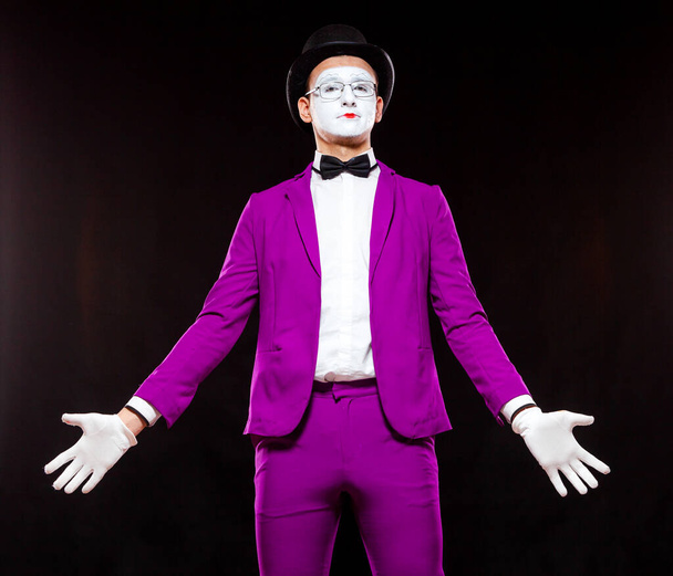 Портрет мужчины-пантомимы, изолированный на черном фоне. Молодой человек в фиолетовом костюме стоит прямо с распростертыми объятьями
 - Фото, изображение