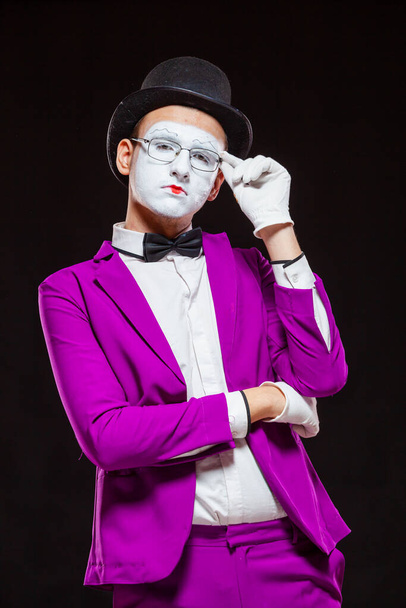 Portrait of male mime artist, isolated on black background. Young man in purple suit is touching his chin with a thoughtful look. Symbol of reflection, criticism - Photo, Image