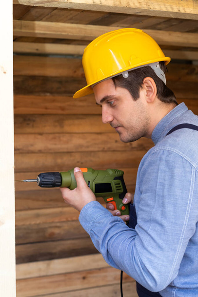 Portrait of a handsome young craftsman with yellow hard hat holding an electric drill  - Fotoğraf, Görsel