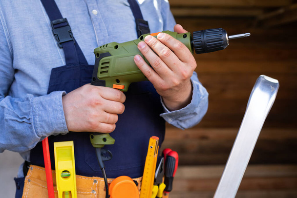 Picture of an electric drill in young man's hands, wooden background outdoor - Foto, afbeelding