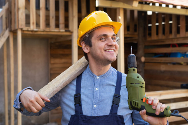 Portrait of a handsome young craftsman with yellow hard hat holding an electric drill  - Zdjęcie, obraz