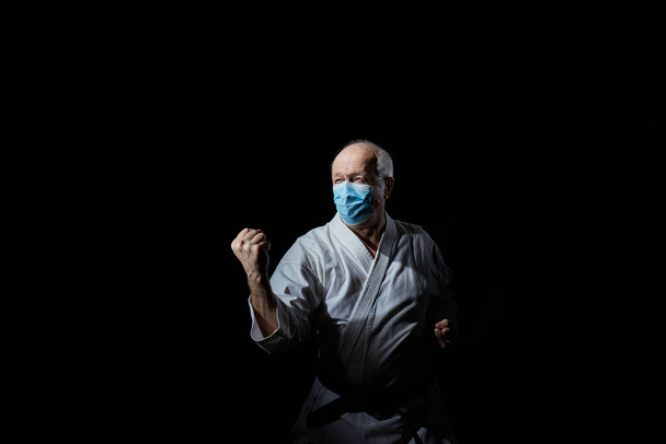 В медицинской маске старый спортсмен тренирует блок рукой на черном фоне.
 - Фото, изображение