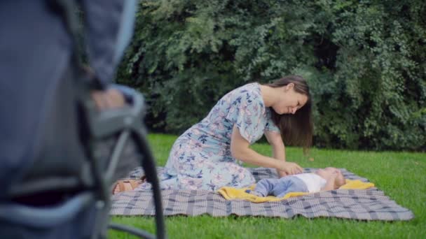Young brunette woman with four-month baby boy sitting on the plaid in the park. Mother smiles at the baby and talks to him. Against the background of a stroller. Slow motion of the camera, 100 fps. - Footage, Video