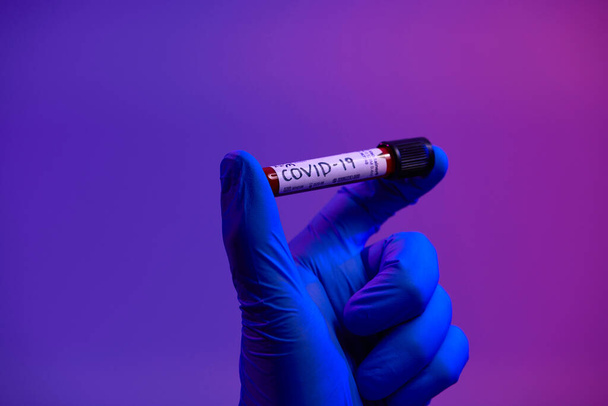 Coronavirus, Doctor holding positive covid-19 virus Blood Sample test tube. Wearing biohazard epidemic Protective mask, suit and glows neon light background - Foto, Imagen