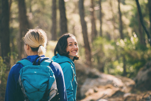 Two girls with backpacks walk in the woods. Hiking in the mountains. Girlfriends spend time together in nature. Women walk in the park. - Foto, imagen