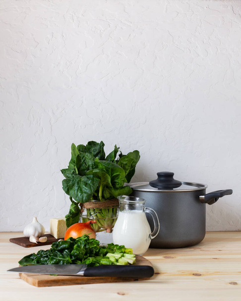 Ingredients for soup puree with raw vegetables, herbs and a pan on light background. Spinach leaves, onion, garlic, butter and milk on wooden table. Cutting spinach with a knife. Vertical, copy space - 写真・画像