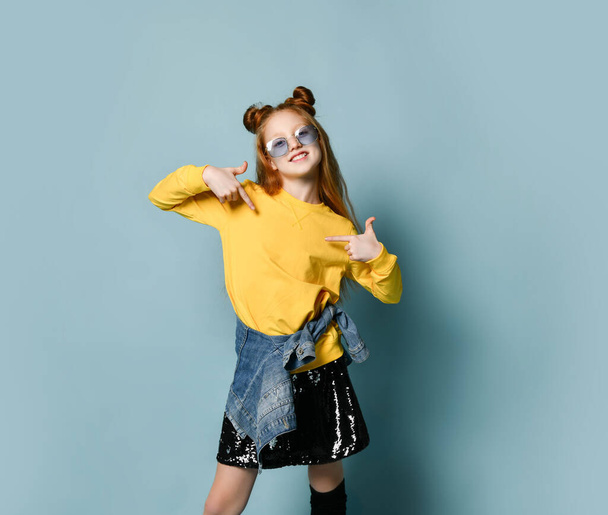 Cool teenager girl with long red hair is pointing fingers of both hands at free copy space on her yellow sweatshirt - Photo, Image