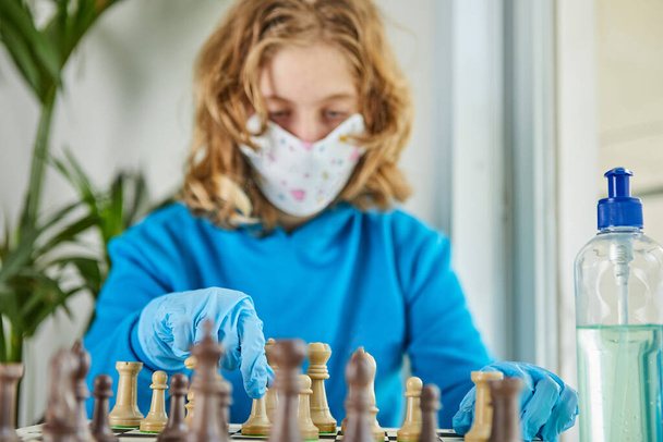 検疫コロナウイルスの生活:検疫covid-19中に自宅で子供たちのためのゲームや活動。仮面と手袋をした10代の少女がチェスをする。選択的焦点. - 写真・画像