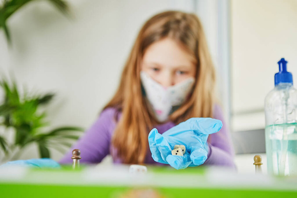 検疫コロナウイルスの生活:検疫covid-19中に自宅で子供たちのためのゲームや活動。マスクと手袋のかわいい女の子はサイコロとチップでゲームをプレイします。選択的焦点. - 写真・画像